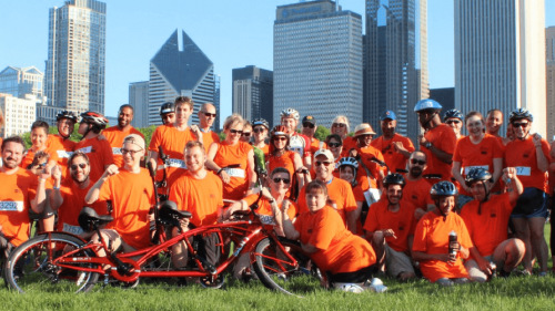 Large group photo outdoors with cityline in the background while wearing orange "Bike the Ride" t-shirts and posing with bikes