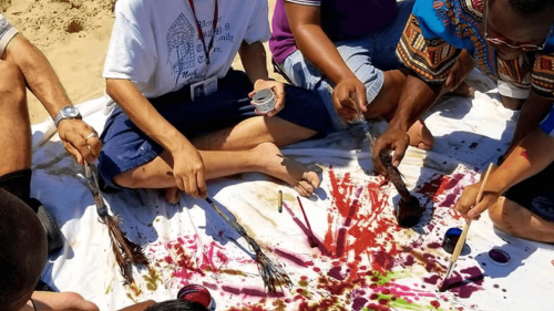 Group of men sitting on a blanket that's placed on the sand and painting