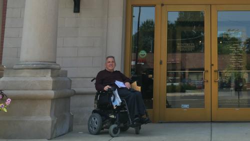 a man sits in an electric wheelchair outside the entrance of a bank with a stone facade