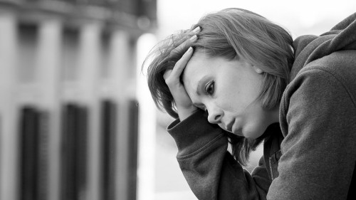 The black and white image shows a person leaning against a wall, looking down with a somber expression. They are wearing a hooded sweatshirt, with the hood down. Their hand is resting on the side of their head, fingers threaded through their hair. The background is blurred, suggesting an urban setting with tall buildings. The grayscale tones emphasize the solemn mood of the scene.