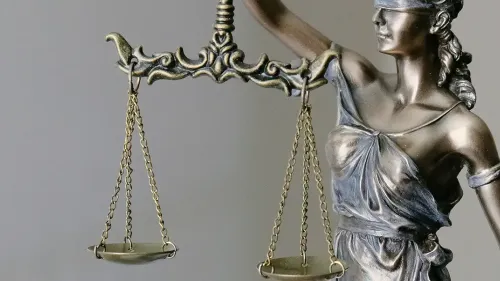 Metallic statue of Lady Justice in profile, holding balanced scales of justice in both hands, wearing a crown and classical robes against a neutral gray background.