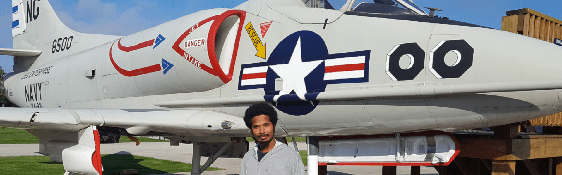 Young black man wearing a gray hoodie standing in front of a jet and giving a thumbs up while smiling