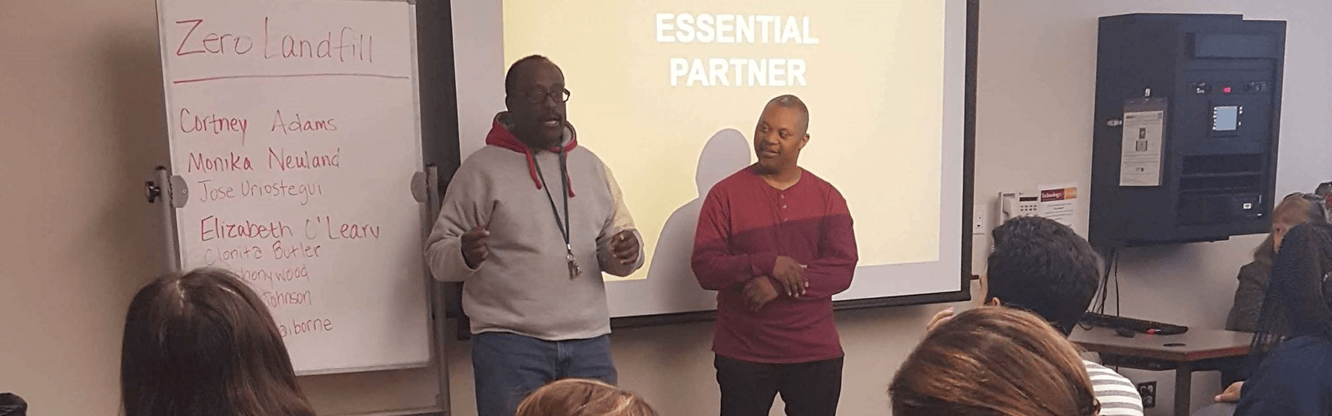 Teacher and student stranding in front of a class by the projection screen and white board