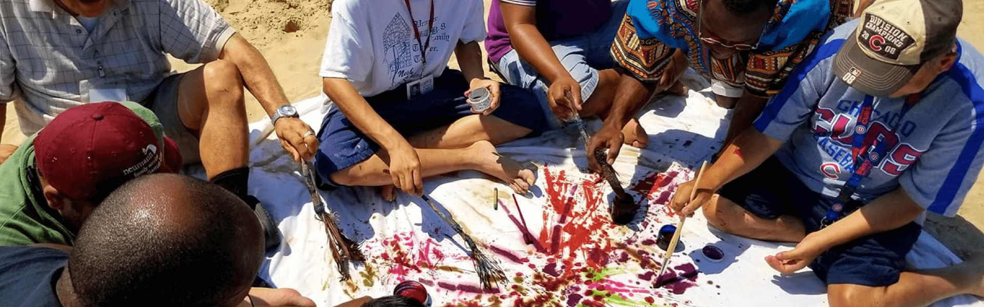 Group of men sitting on a blanket that's placed on the sand and painting