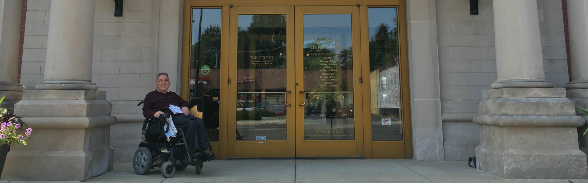 a man sits in an electric wheelchair outside the entrance of a bank with a stone facade