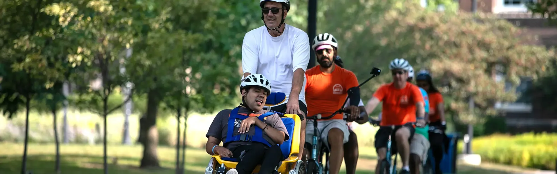 An adult cyclist leads a group ride while towing a child in a yellow cargo bike trailer on a sunny park path. Multiple cyclists in orange jerseys follow behind on the tree-lined, paved route.