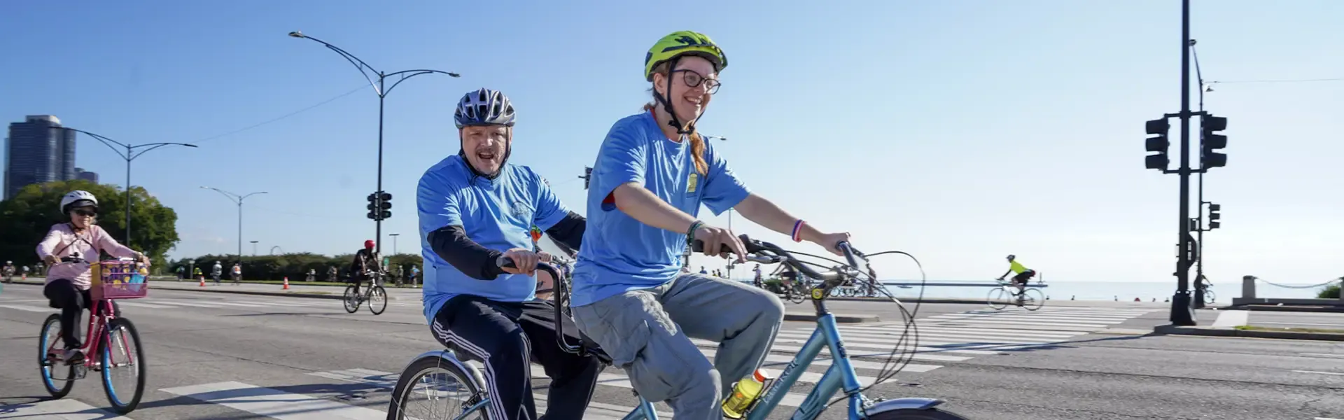Two cyclists in blue shirts riding a blue tandem bicycle on a city street with downtown high-rise buildings in the background, traffic signals, and other cyclists visible.