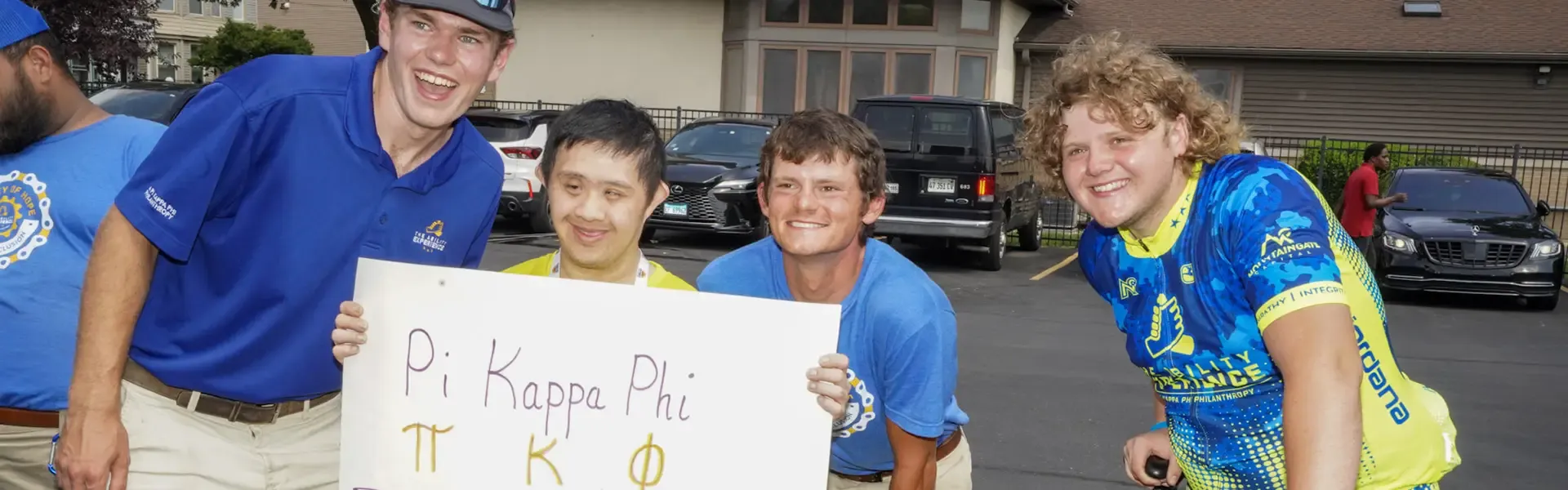 Group of four cyclists in blue and yellow athletic wear posing at a parking lot holding a banner that reads 'Pi Kappa Phi ΠΚΦ ROCKS,' displaying camaraderie at a cycling event.