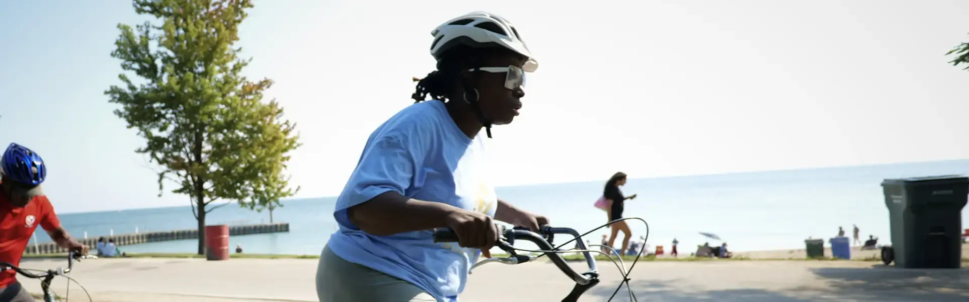 Cyclist in a blue shirt riding a turquoise bicycle on a waterfront recreational path with calm blue water, a tree, and beachgoers visible in the background.