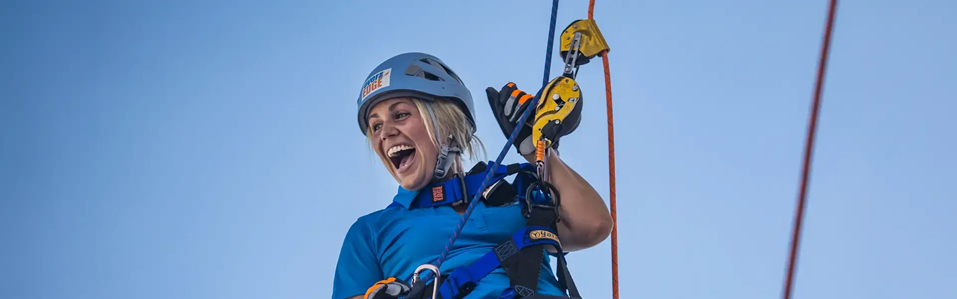 Person in blue safety gear and helmet smiling while rappelling outdoors, holding yellow and orange climbing carabiners attached to secured rope lines.