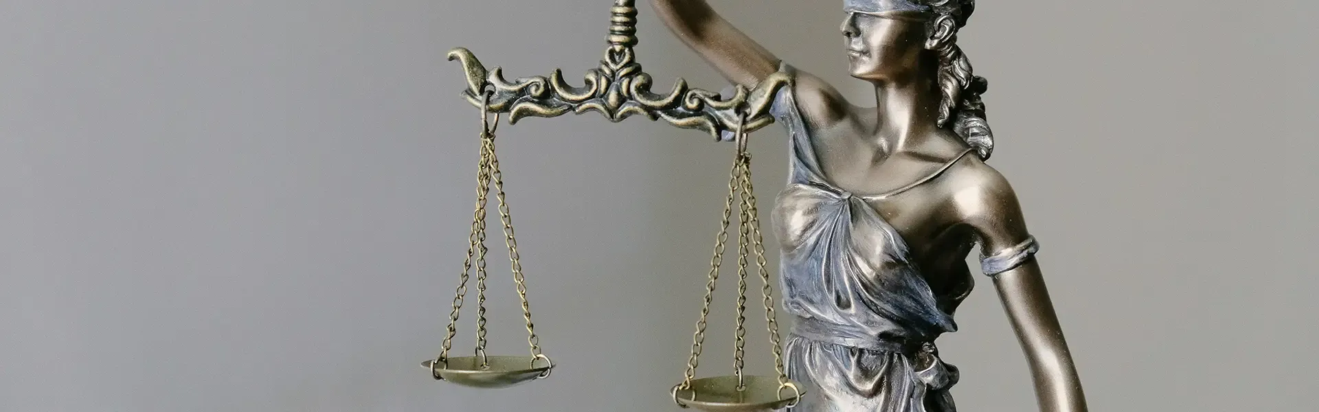 Metallic statue of Lady Justice in profile, holding balanced scales of justice in both hands, wearing a crown and classical robes against a neutral gray background.
