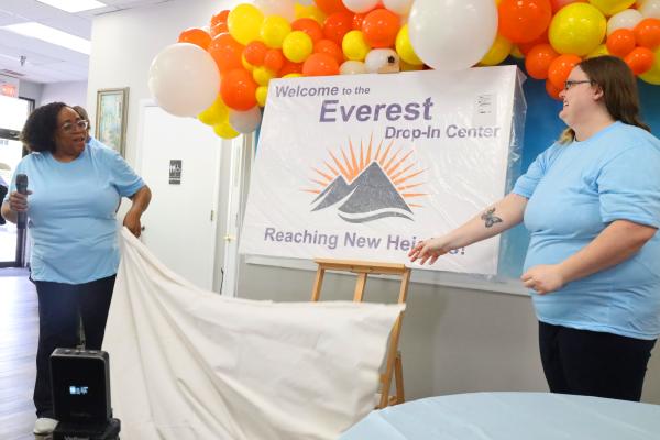Two people in blue shirts unveil a sign, with orange, yellow, and white balloons displayed above in the background.
