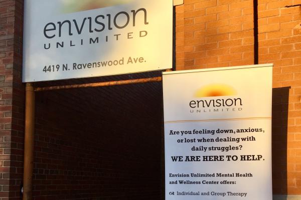 A sunlit doorway on a red brick wall with white signage featuring a yellow and orange sunburst logo reading 'Envision Unlimited' with details about mental health offerings.