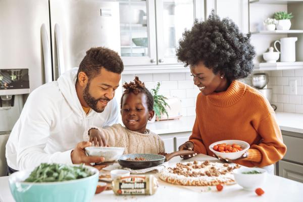 A family prepares a meal together in a kitchen; a person in a white hoodie and a person in an orange turtleneck holding a bowl of ingredients stand on either side of a child who reaches into a bowl, with various ingredients on the counter in front of them.
