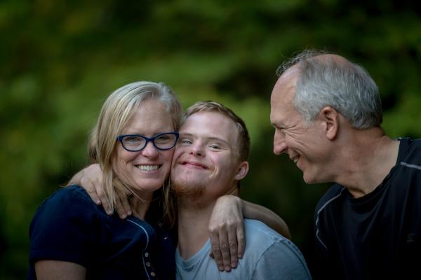 Three smiling people stand in front of a green background; the person on the left wears glasses, the person in the middle wears a blue top, and the person on the right wears a dark top.