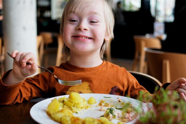 A smiling child in an orange t-shirt holds a fork over a half-finished meal.