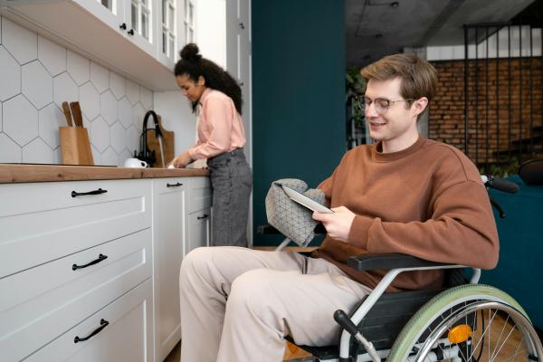 Two people work together in a white kitchen with wood accents; a person in a wheelchair wearing a brown sweater and glasses dries a dish in the foreground, while another in a pink top and dark jeans stands at the sink in the background.