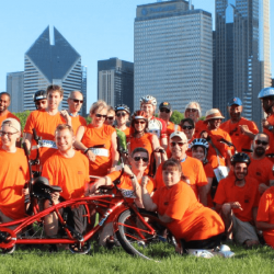 Large group photo outdoors with cityline in the background while wearing orange "Bike the Ride" t-shirts and posing with bikes