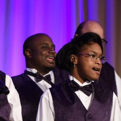 Group of people dressed up in purple vest and bow ties with white button down shirts singing on stage