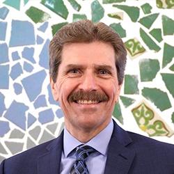 Mark smiling and wearing a blue suit and tie while in front of a colorful background