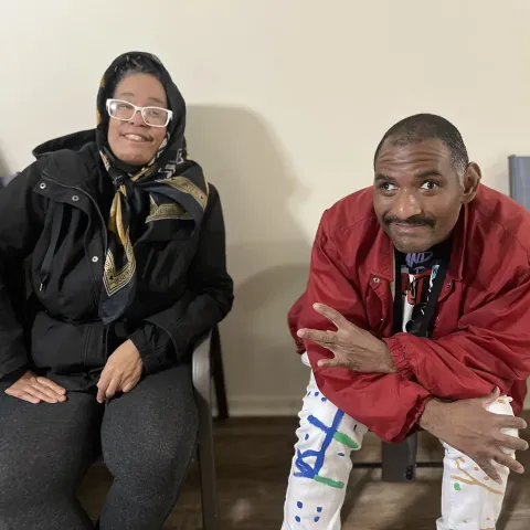 Two people seated indoors, one in a headscarf and black jacket, the other in a red jacket with colorful pants.