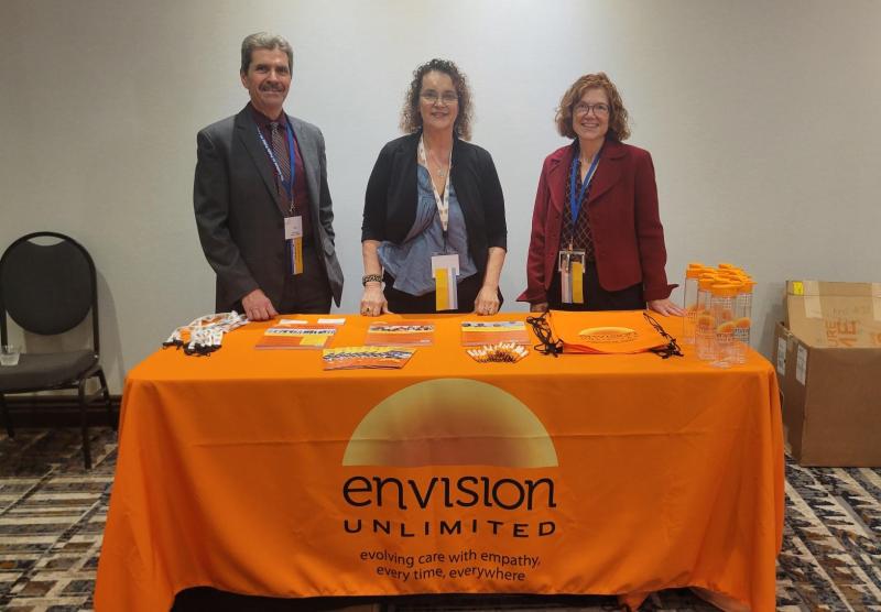 Three staff members stand at a booth with a bright orange tablecloth at a conference.