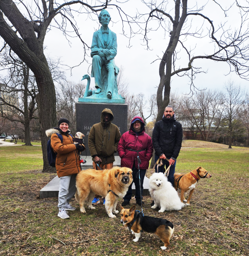 A group of people walking dogs stand in front of a statue in a park.