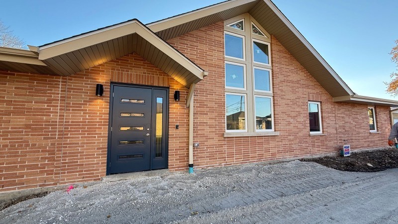 The image depicts a single-story house with a red brick facade and a steep, gabled roof. The front entrance features a modern dark-colored door with four narrow horizontal windows. Two wall-mounted lights flank the door, casting a warm glow on the brick surface. To the right of the door, there are four large vertical windows arranged in a column, providing a symmetrical look. The roof overhangs slightly, providing shade and adding emphasis to the architectural design. The ground in front of the house consis