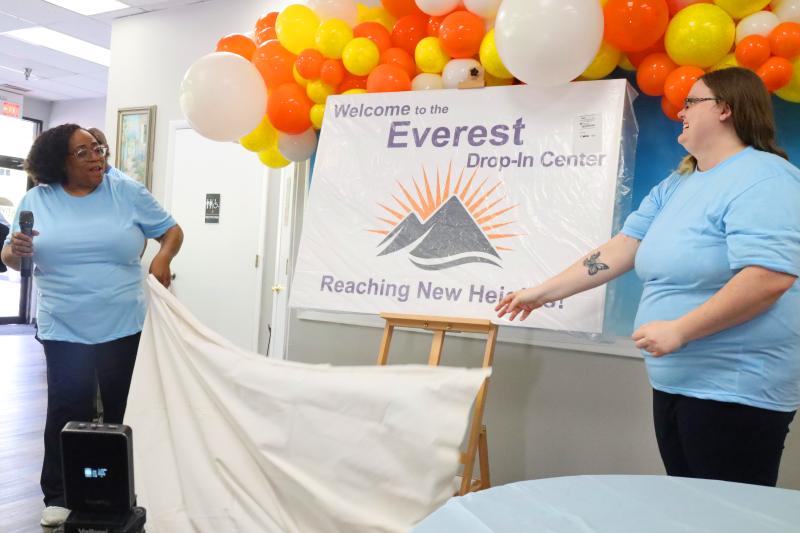 Two people in blue shirts unveil a sign, with orange, yellow, and white balloons displayed above in the background.