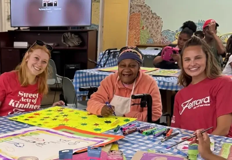 Three women smiling at an art table covered with markers and paintings.
