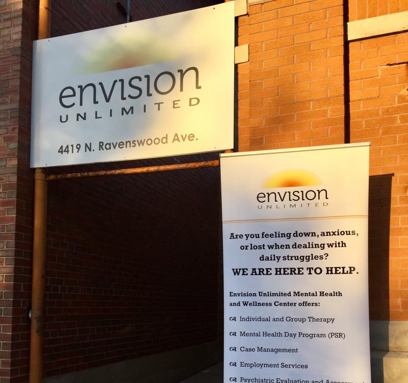 A sunlit doorway on a red brick wall with white signage featuring a yellow and orange sunburst logo reading 'Envision Unlimited' with details about mental health offerings.