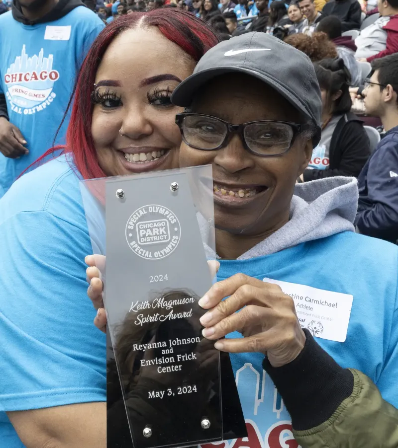 Two individuals smiling and holding an award plaque at a public event.