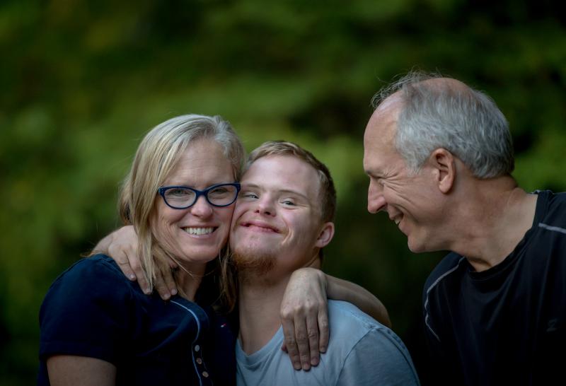 Three smiling people stand in front of a green background; the person on the left wears glasses, the person in the middle wears a blue top, and the person on the right wears a dark top.