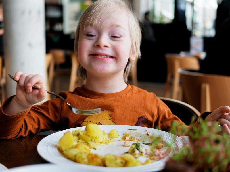 A smiling child in an orange t-shirt holds a fork over a half-finished meal.