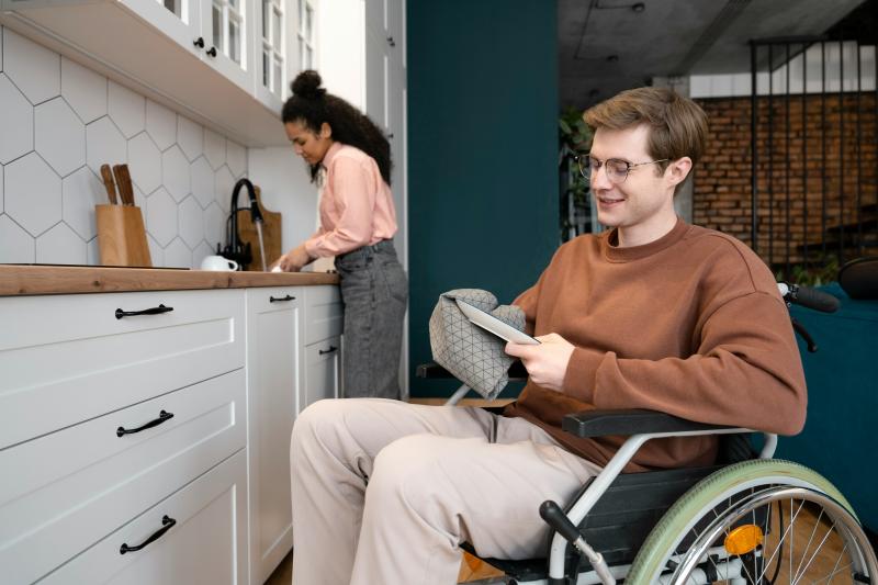 Two people work together in a white kitchen with wood accents; a person in a wheelchair wearing a brown sweater and glasses dries a dish in the foreground, while another in a pink top and dark jeans stands at the sink in the background.