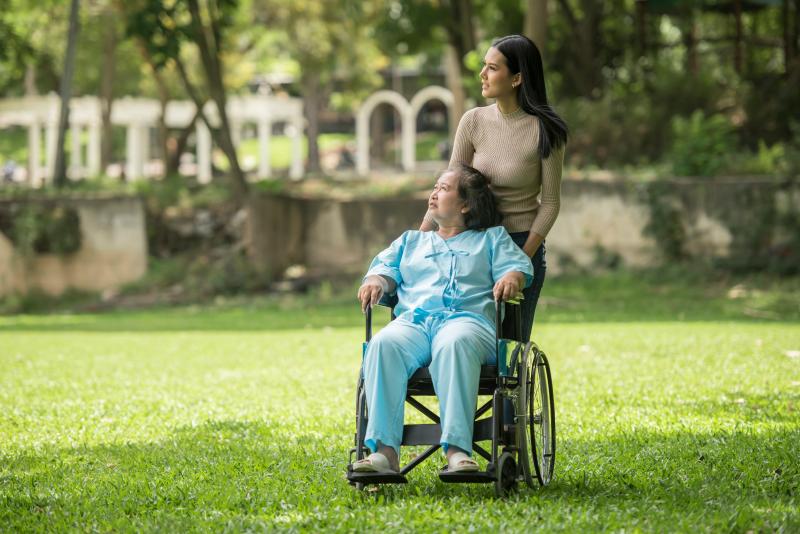 A person in a tan sweater walks behind a wheelchair user in a blue outfit on a sunny day outside, surrounded by green grass, both looking to the left.