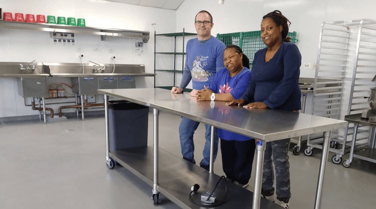 Three people standing at a steel counter in a commercial kitchen and smiling at the camera