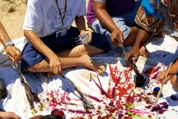 Group of men sitting on a blanket that's placed on the sand and painting