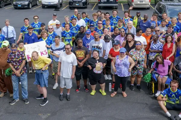 Large diverse group of over 100 people wearing blue 'Journey of Hope' shirts gathered in a parking lot, smiling and waving at the camera.