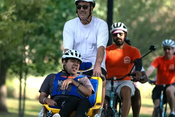 An adult cyclist leads a group ride while towing a child in a yellow cargo bike trailer on a sunny park path. Multiple cyclists in orange jerseys follow behind on the tree-lined, paved route.