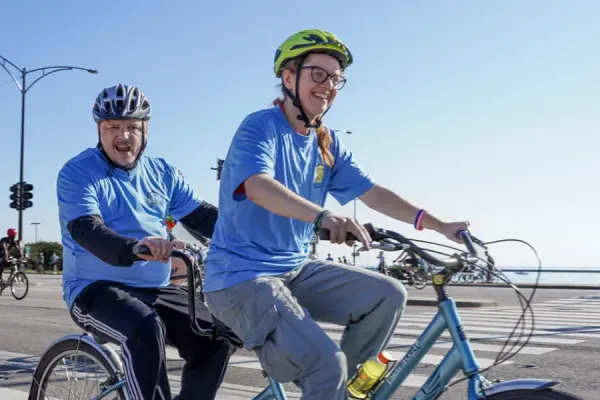Two cyclists in blue shirts riding a blue tandem bicycle on a city street with downtown high-rise buildings in the background, traffic signals, and other cyclists visible.
