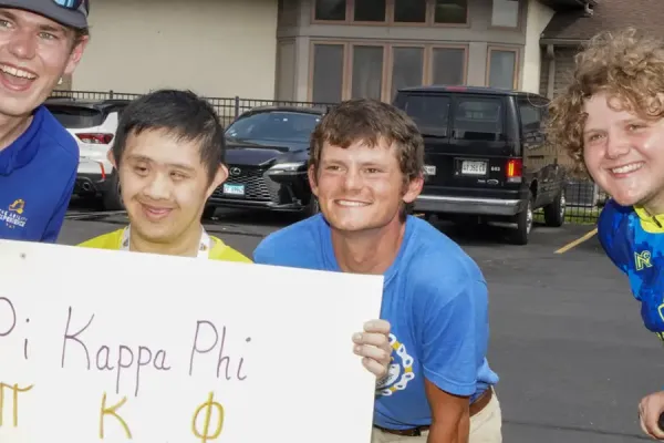 Group of four cyclists in blue and yellow athletic wear posing at a parking lot holding a banner that reads 'Pi Kappa Phi ΠΚΦ ROCKS,' displaying camaraderie at a cycling event.