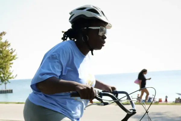 Cyclist in a blue shirt riding a turquoise bicycle on a waterfront recreational path with calm blue water, a tree, and beachgoers visible in the background.