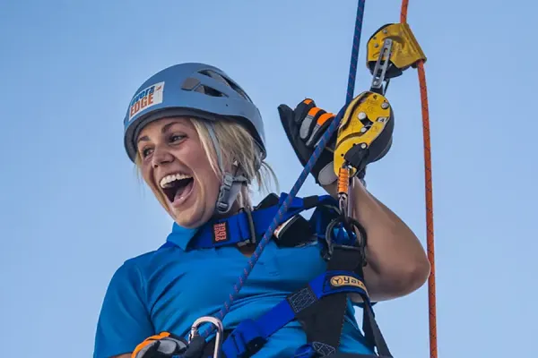 Person in blue safety gear and helmet smiling while rappelling outdoors, holding yellow and orange climbing carabiners attached to secured rope lines.