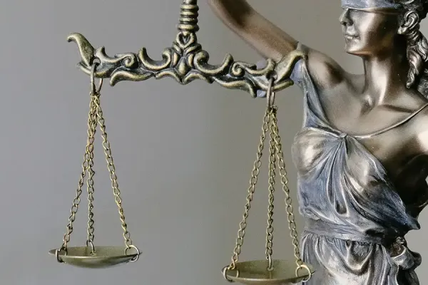 Metallic statue of Lady Justice in profile, holding balanced scales of justice in both hands, wearing a crown and classical robes against a neutral gray background.